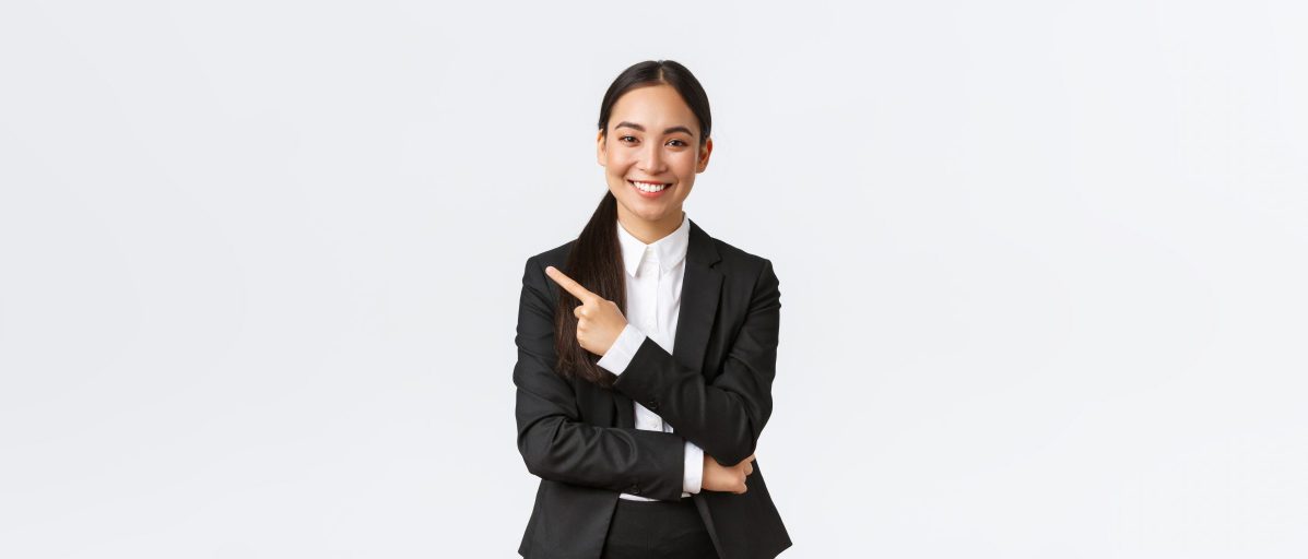 Happy professional asian female manager, businesswoman in suit showing announcement, smiling and pointing finger left at product or project banner, standing white background.
